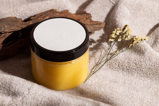 Jar of yellow skincare cream on a towel with wood and dried flowers, ideal for spa themes.
